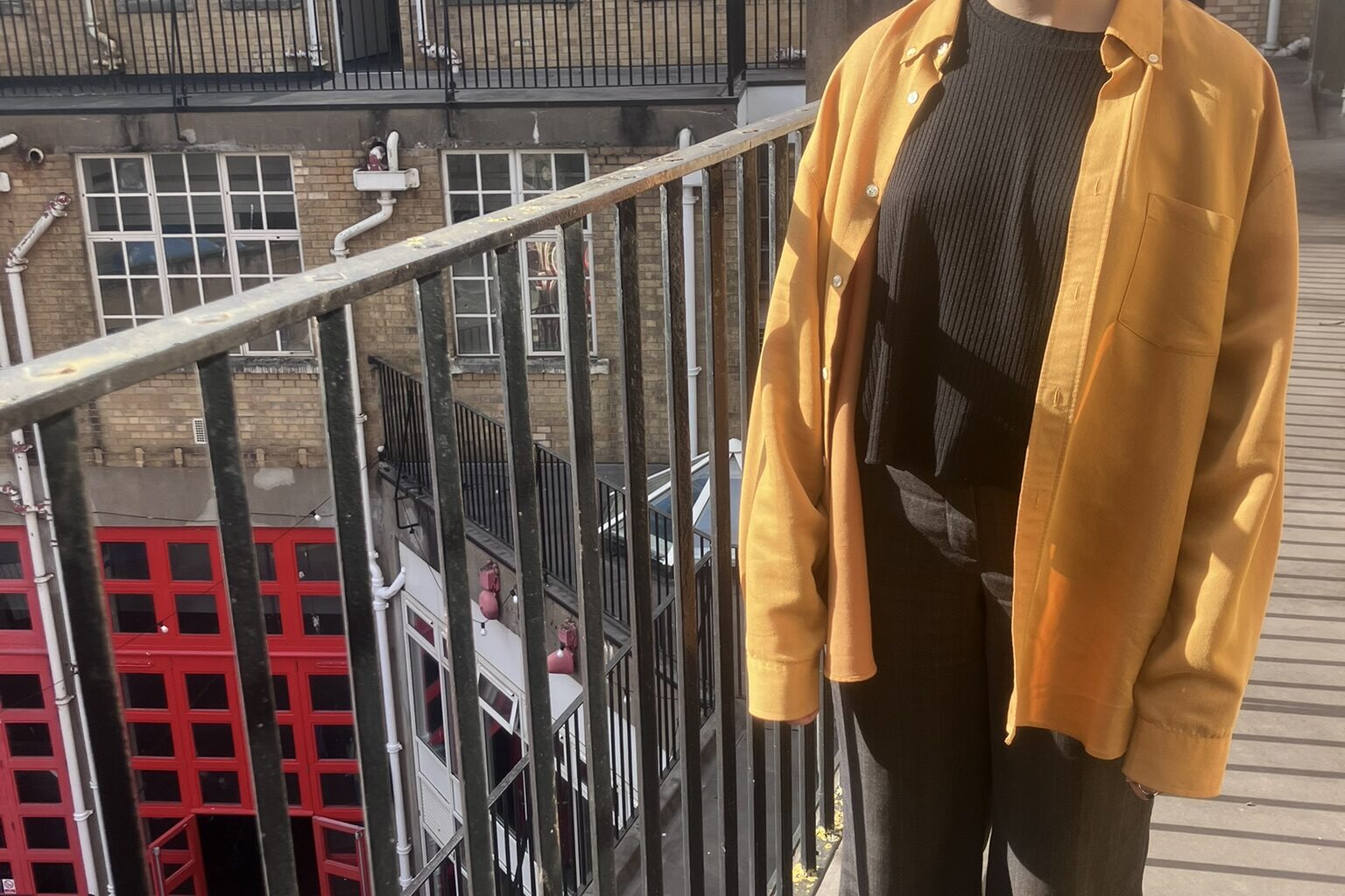 A woman and standing and smiling on a balcony, wearing a yellow jacket and black trousers. A bright red tour is on the ground floor.