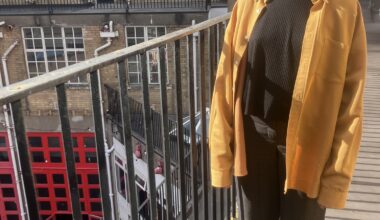 A woman and standing and smiling on a balcony, wearing a yellow jacket and black trousers. A bright red tour is on the ground floor.