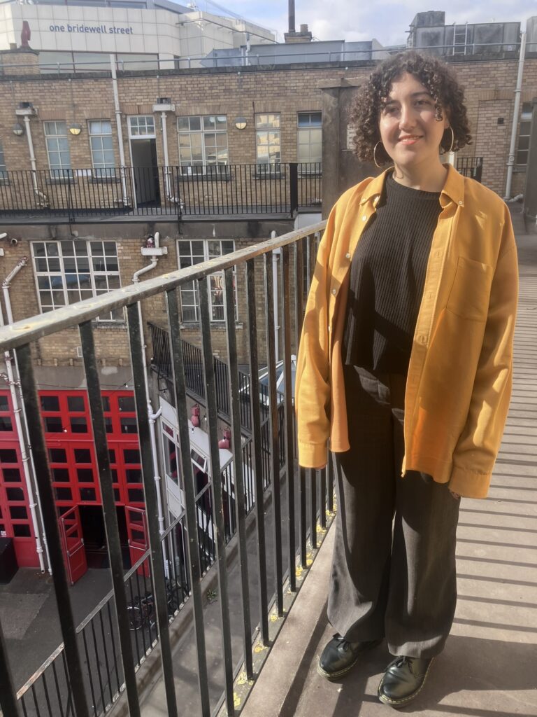 A woman and standing and smiling on a balcony, wearing a yellow jacket and black trousers. A bright red tour is on the ground floor.