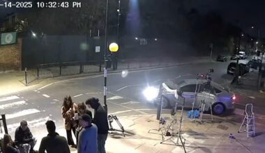 A nighttime street scene shows a group of people gathered on a sidewalk near film equipment. A silver car is parked by a zebra crossing, and bright lights illuminate the area. The timestamp reads “04-14-2025 10:32:43 PM.”.