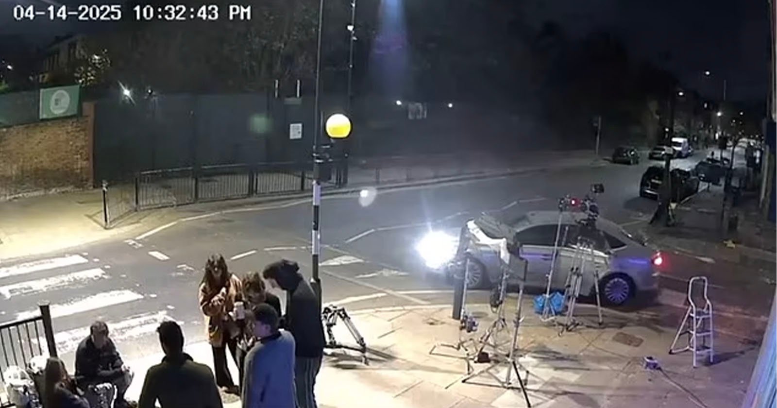 A nighttime street scene shows a group of people gathered on a sidewalk near film equipment. A silver car is parked by a zebra crossing, and bright lights illuminate the area. The timestamp reads “04-14-2025 10:32:43 PM.”.