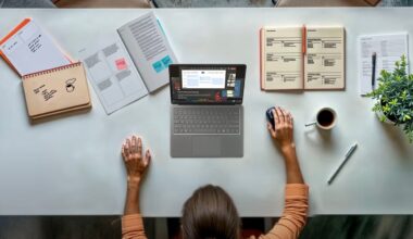 A person sits at a tidy desk with a laptop, notebooks, papers, a pen, a coffee cup, and a small potted plant, working with one hand on the mouse and the other resting on the desk.
