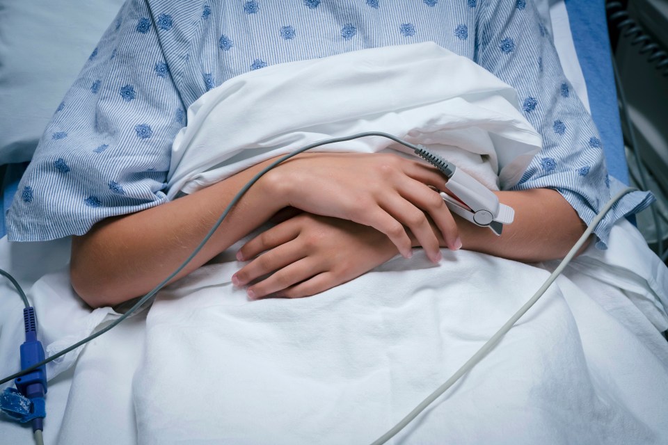 A child's hands clasped on their stomach in a hospital bed, wearing a pulse oximeter.