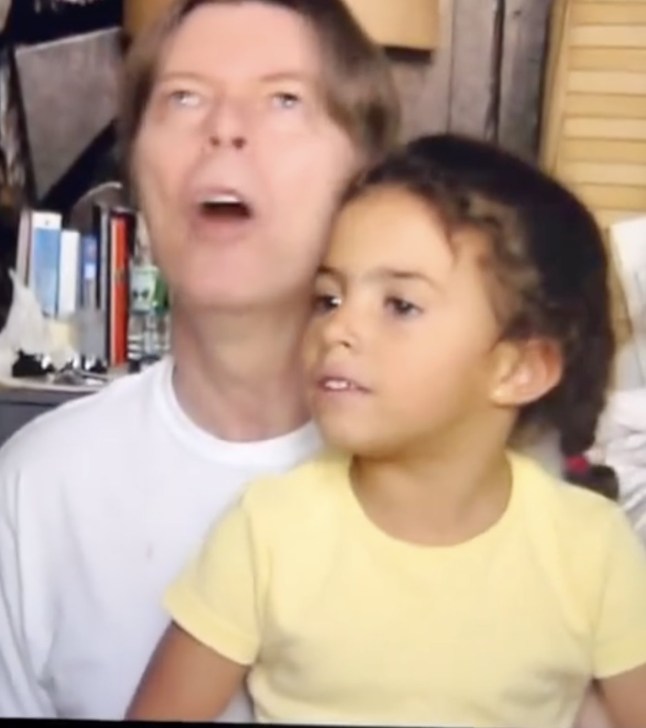 David Bowie sings as his young daughter sits on his lap in yellow top