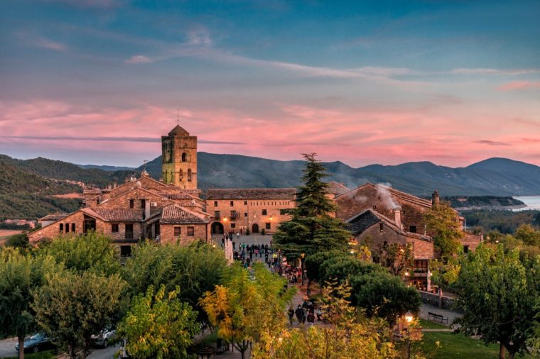 Groups of people, of all sexes and ages, stroll through the square, in the background the tower of the church of Santa Maria at sunset Characterized by its medieval town planning with castle and walls. Ainsa, Huesca, Aragon, Spain. November 3, 2018