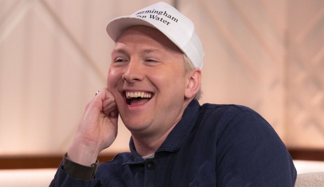 Joe Lycett laughs while wearing a white baseball cap with the slogan Birmingham Tap Water' written on it