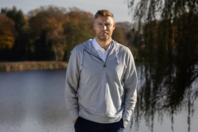 Freddie Flintoff standing in front of a lake and trees. He's wearing a grey jumper with his hands in his pockets, and a white shirt underneath.