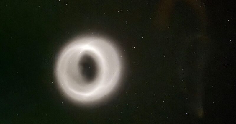 A glowing, ring-shaped phenomenon appears in the night sky, surrounded by scattered stars. The ring emits a soft white light, creating a striking contrast against the dark background.