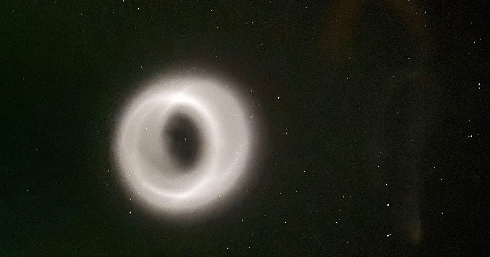 A glowing, ring-shaped phenomenon appears in the night sky, surrounded by scattered stars. The ring emits a soft white light, creating a striking contrast against the dark background.