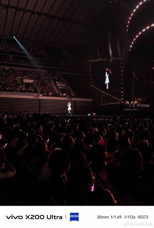 A concert hall filled with an audience, some holding light sticks. A performer on stage is wearing a white outfit. The stage is illuminated with bright lights and a large screen shows the performer. Camera details are noted at the bottom.