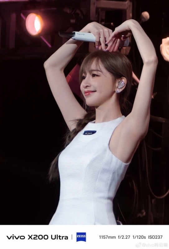 A woman in a white sleeveless dress is on stage making a heart shape with her arms above her head. She is smiling, with her hair tied back, and there are stage lights and equipment in the background.