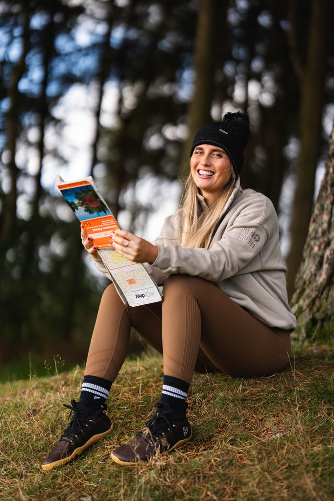 Woman in leggings and a fleece sitting on a mossy hill smiling and looking at a map