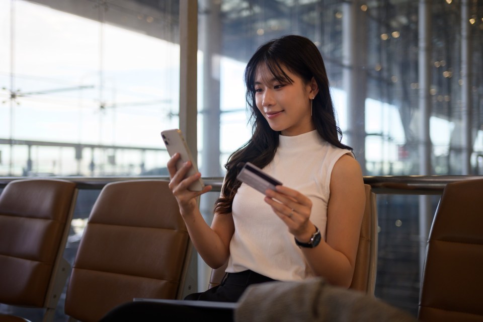 Young asian woman sitting in an airport waiting area, using her smartphone and credit card for an online purchase, enjoying the convenience of digital shopping while traveling