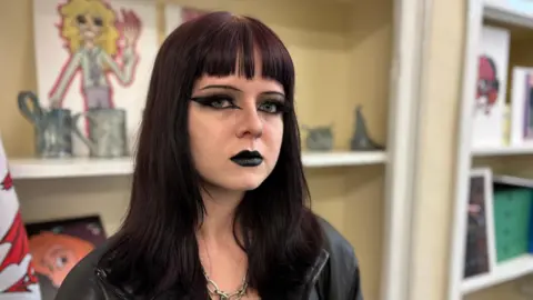 BBC Lili, with long black hair, dark eye make-up and black lipstick, is sitting in a black jacket in front of some shelves with various items on them.