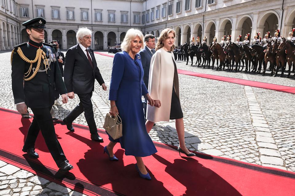 Queen Camilla wearing a blue coat walking down a red carpet in Italy with soldiers standing in a row behind her