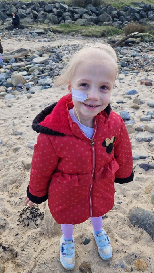 A young girl with a nasal cannula stands on a beach.
