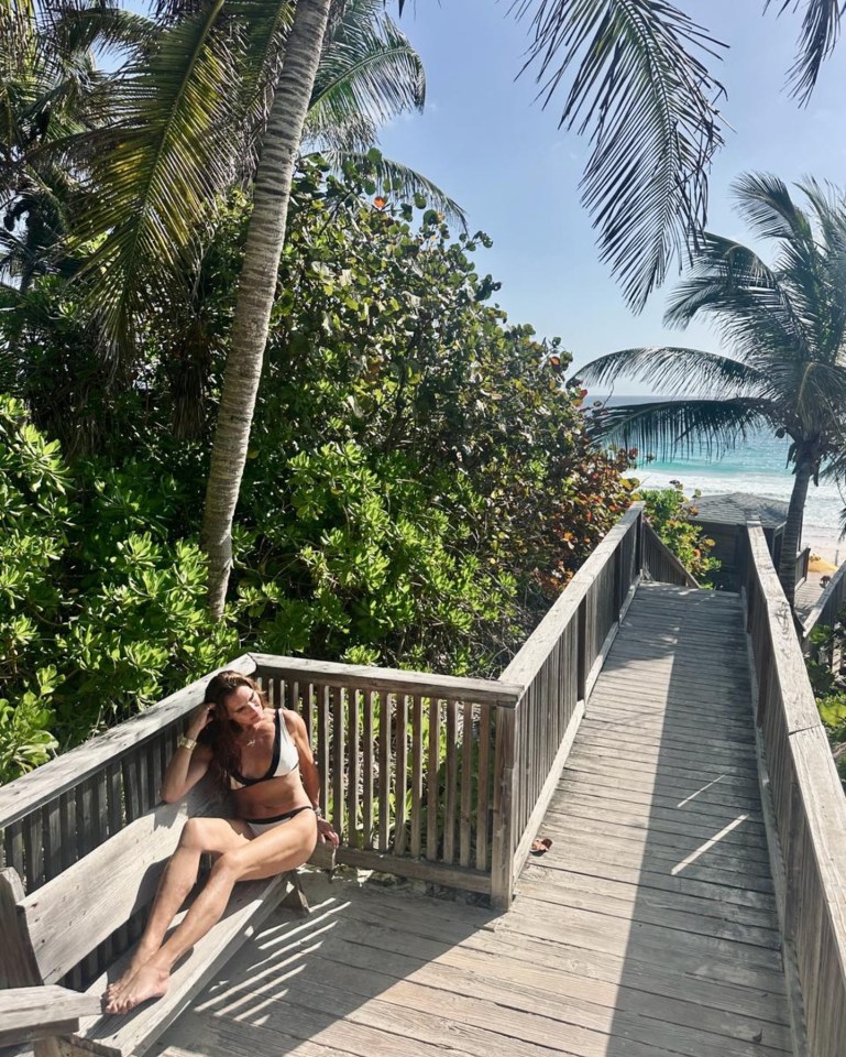 Brooke Shields in a white bikini sitting on a bench overlooking a beach.