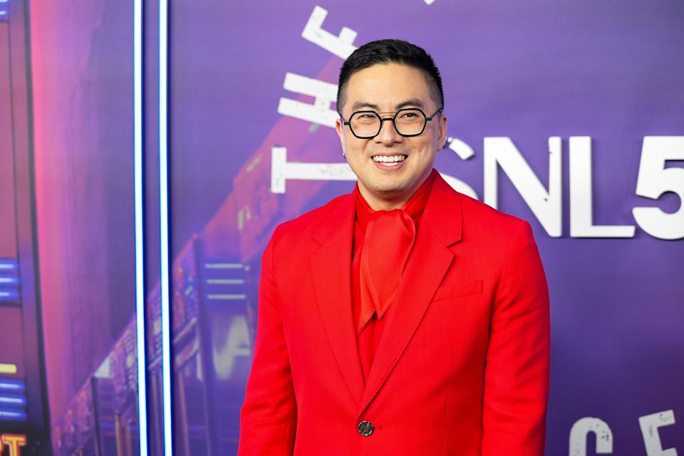 Bowen Yang in bold suit with scarf, standing in front of "SNL" backdrop, smiling