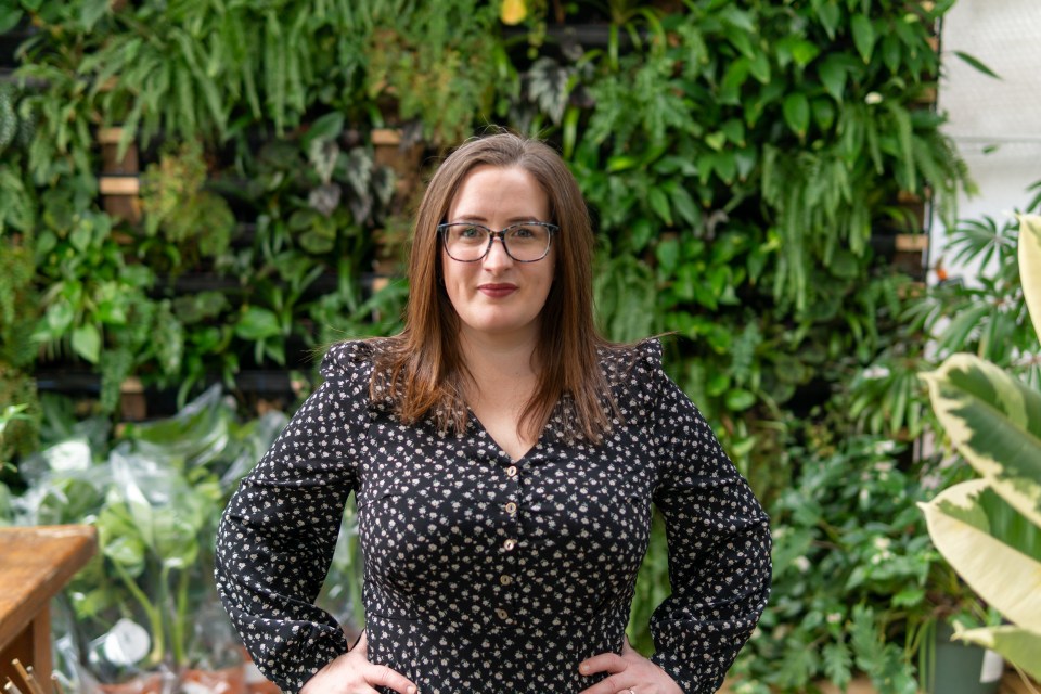 Portrait of a woman wearing glasses and a floral dress, standing in front of a wall of plants.