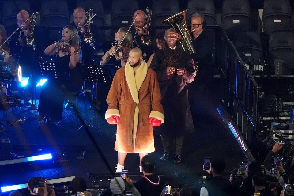 Chris Eubank Jr. in a brown robe and boxing gloves, standing before an orchestra.