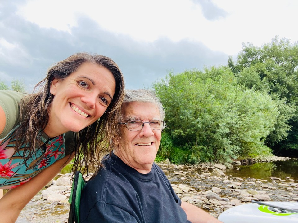 A woman and her father, who has dementia, smile for a photo by a river.