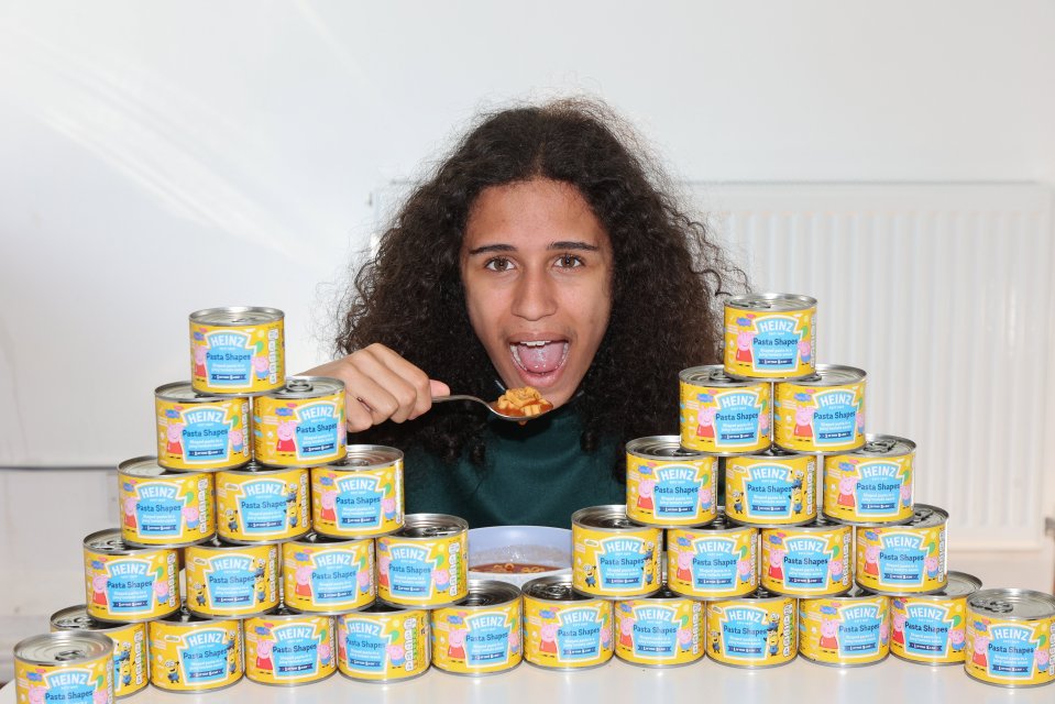 Person eating Heinz Peppa Pig pasta shapes surrounded by many cans of the product.