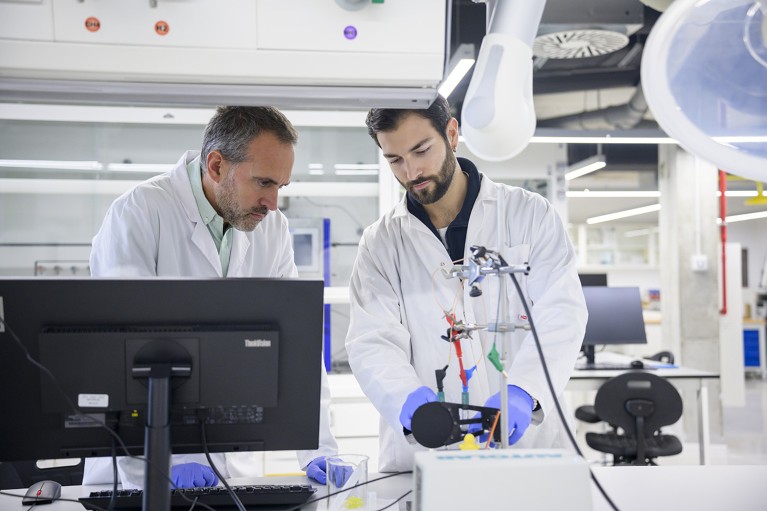 In the foreground on left, a man wearing white coat and purple gloves stands behind a computer screen looking down towards the right at some equipment. To his right, a man wearing white coat and purple gloves stands behind equipment, also looking down towards it.