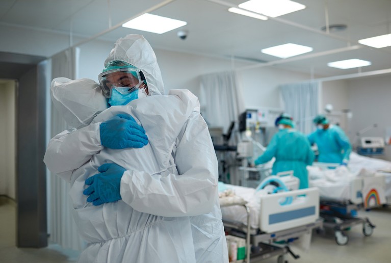 Two people in full personal protective equipment embrace supportively in a hospital ward.