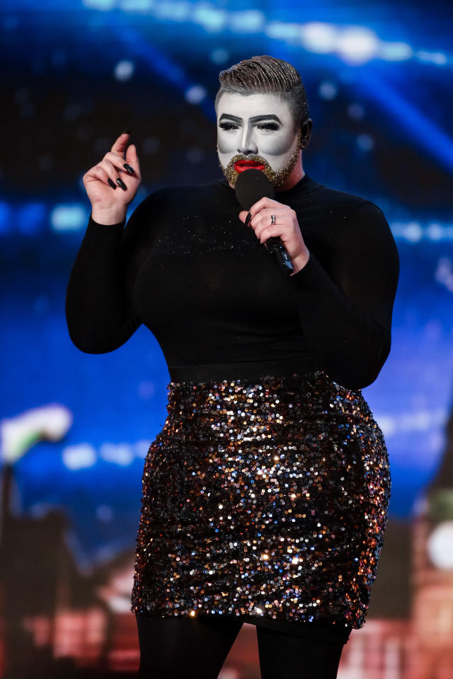 Person in black top and sequined skirt with mime makeup holding a microphone.
