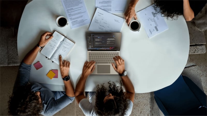 Three people sit around a white table with papers, notebooks, and coffee cups, collaborating while one person types on a laptop displaying charts and code.