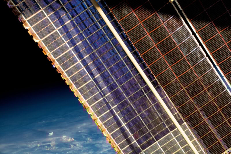 Close-up view of two solar panel arrays from the International Space Station, with Earth’s blue atmosphere and clouds visible in the background against the darkness of space.