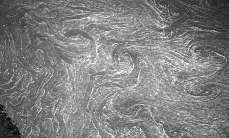 Black and white aerial view of swirling, spiral patterns on a water surface, resembling abstract art or fluid motion; darker area visible in the bottom left corner.