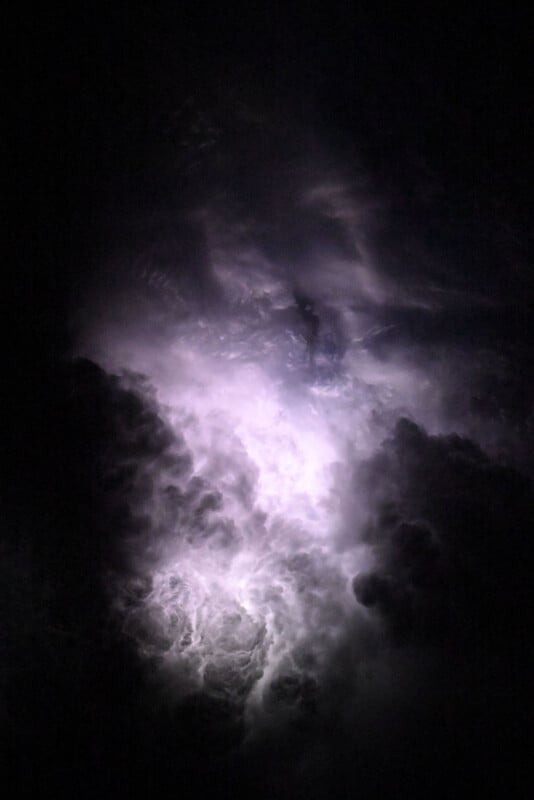 A dramatic, dark sky filled with swirling clouds, illuminated in the center by a bright flash of lightning, creating a glowing, ethereal effect against the night.
