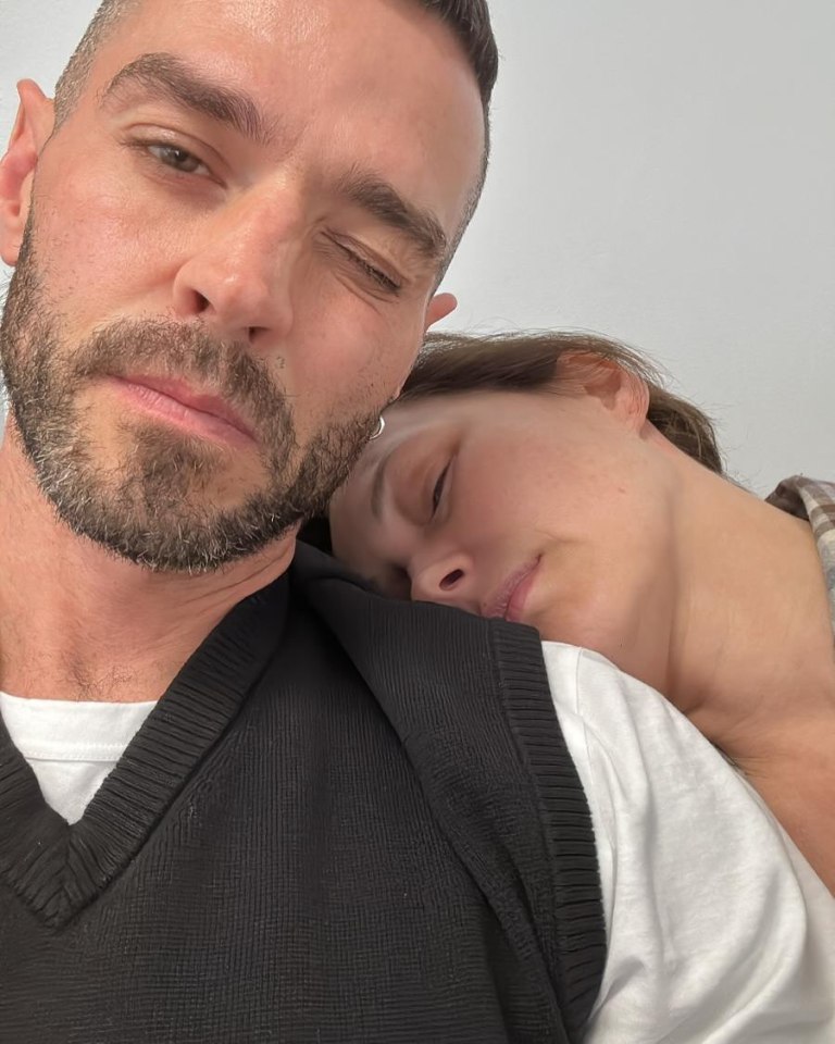 A man with a beard winks at the camera as a woman rests her head on his shoulder.