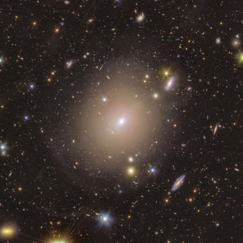 A vibrant image of the Virgo Cluster, showcasing numerous galaxies and stars scattered across the dark expanse of space. The central galaxy is a large elliptical, glowing softly amidst the diverse, luminous specks of distant celestial bodies.