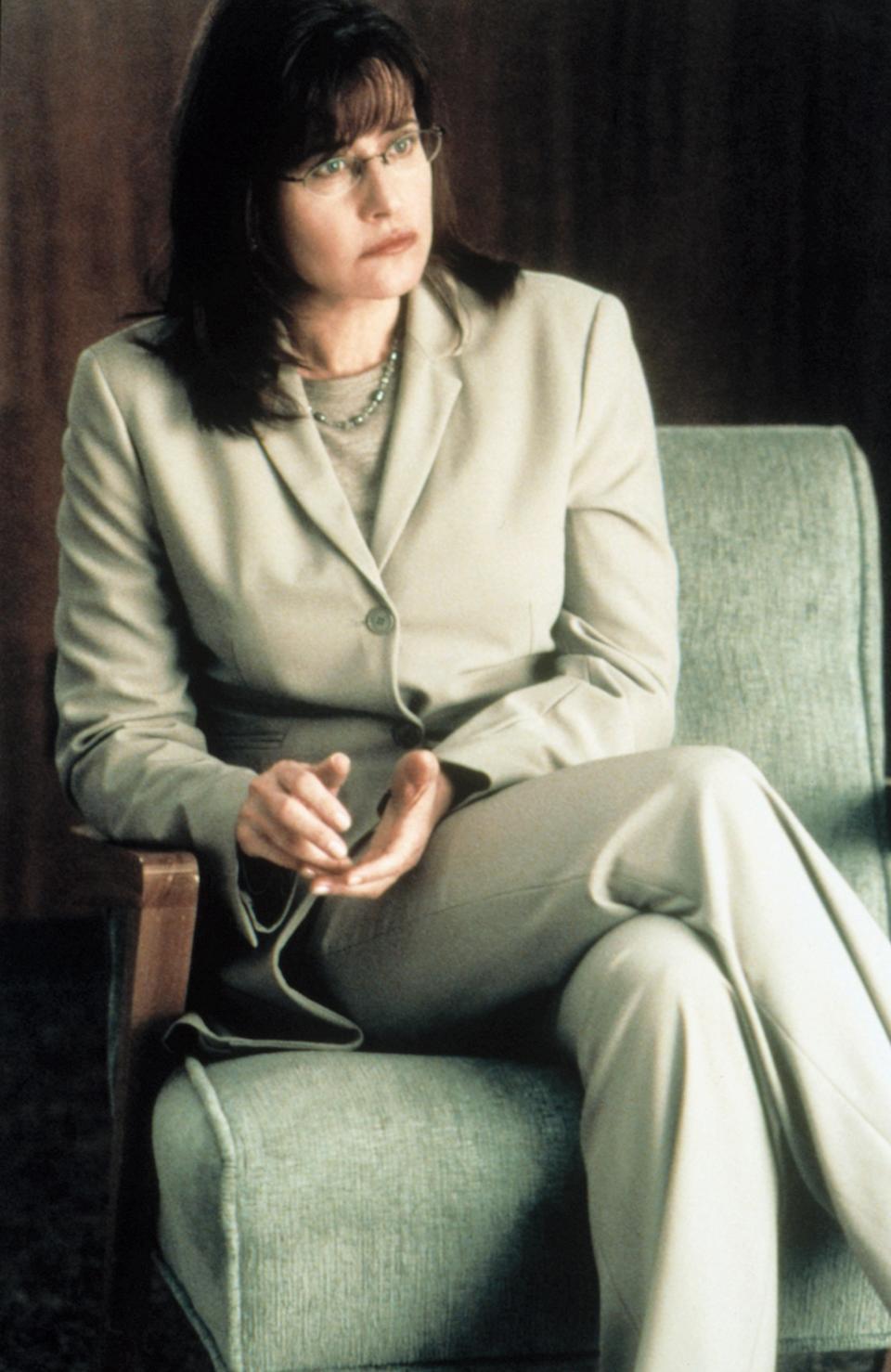 Person in a professional suit sitting thoughtfully in a chair, with glasses and short hair, holding notes