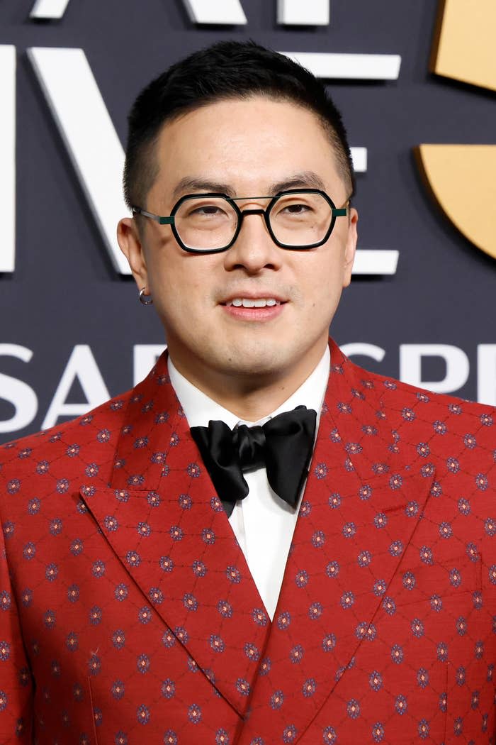 Bowen Yang wearing a patterned suit and round glasses, posed on a formal event backdrop