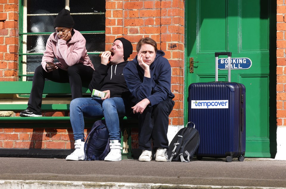 Three people sitting on a bench at a train station, looking frustrated.  A suitcase is nearby.