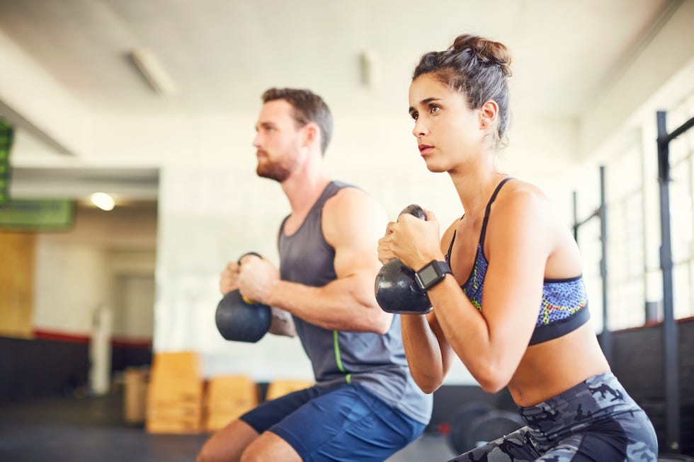 determined female athlete with instructor exercising in health club. they are lifting kettlebells. both are in sports wear.
