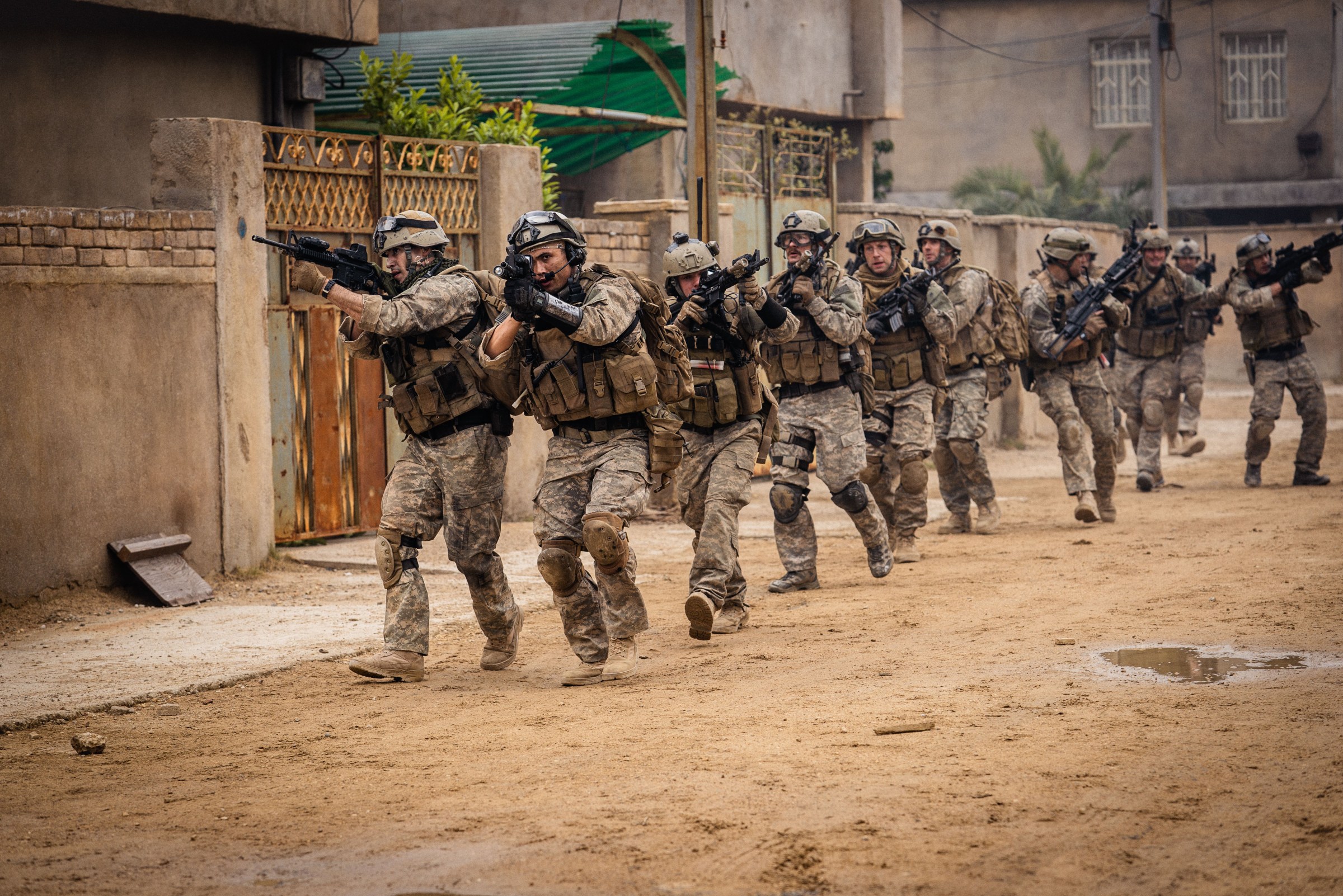Several Navy SEALs walk through a sandy, unpaved Iraq street with their weapons held up in the movie Warfare
