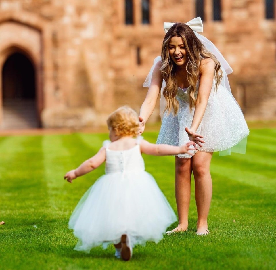A woman in a white dress reaches for a toddler girl in a white dress running on a lawn.