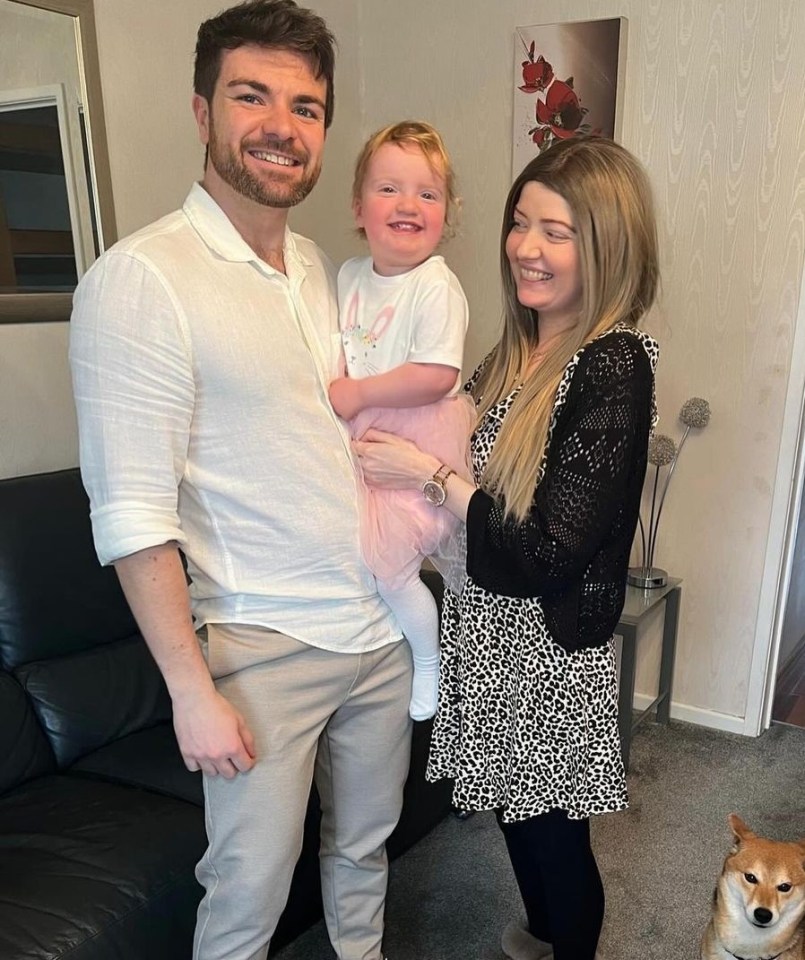 A family portrait: a father holding their toddler daughter, and the mother standing beside them.