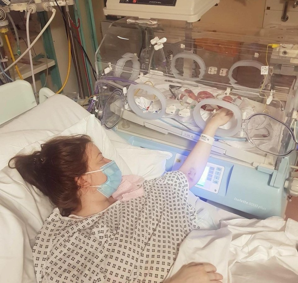 A mother in a hospital bed reaches out to touch her premature baby in an incubator.