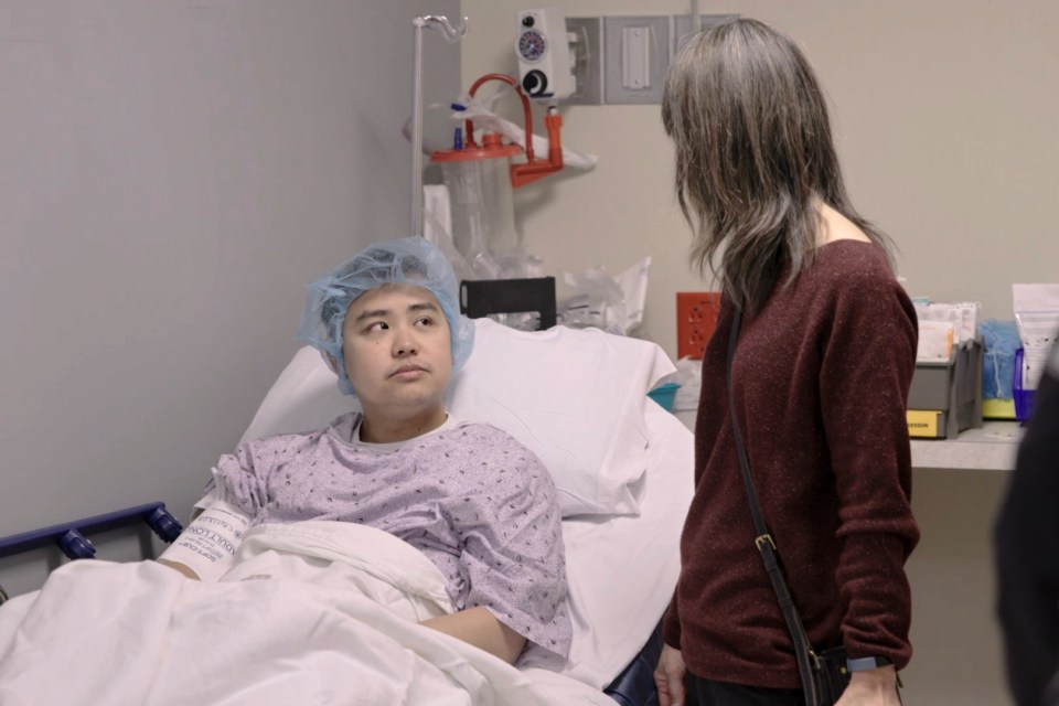 A man in a hospital gown sits in a hospital bed talking to a woman.