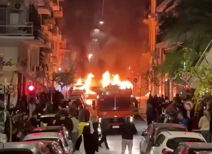 Burning vehicle in city street surrounded by onlookers.