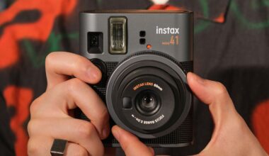A person holds a black Instax Mini 40 instant camera, showing the lens and flash. The camera has a textured body, and the person is wearing a patterned shirt and a ring on their right hand.