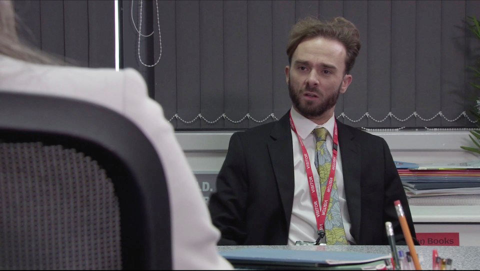 A man in a suit sits at a desk and speaks with someone off-camera.