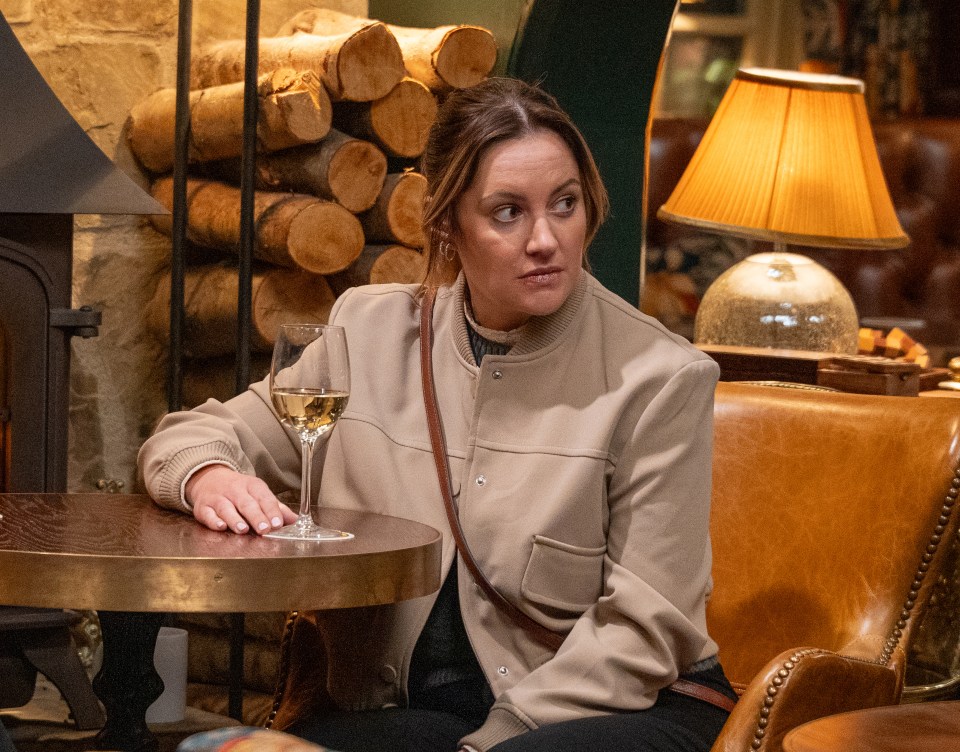 A woman sits at a table in a pub, looking pensive.  She has a glass of white wine in front of her.