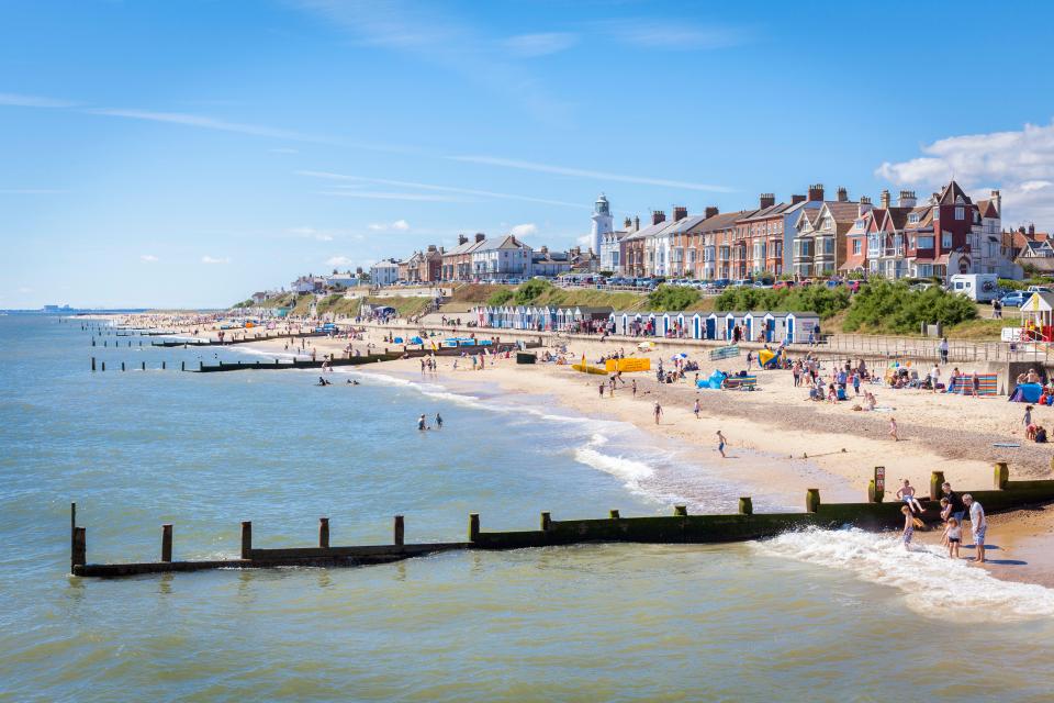 Crowded beach in Southwold, Suffolk, with beach huts and houses along the coast.