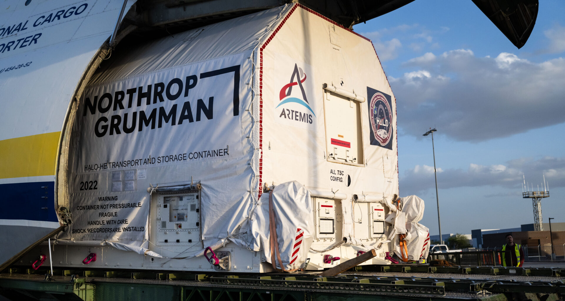 A large cargo aircraft with its nose lifted open reveals a wrapped container being offloaded. The container is marked with the logo for NASA's Artemis campaign and labeled as the HALO transport and storage container. The scene is set at an airport during late afternoon.
